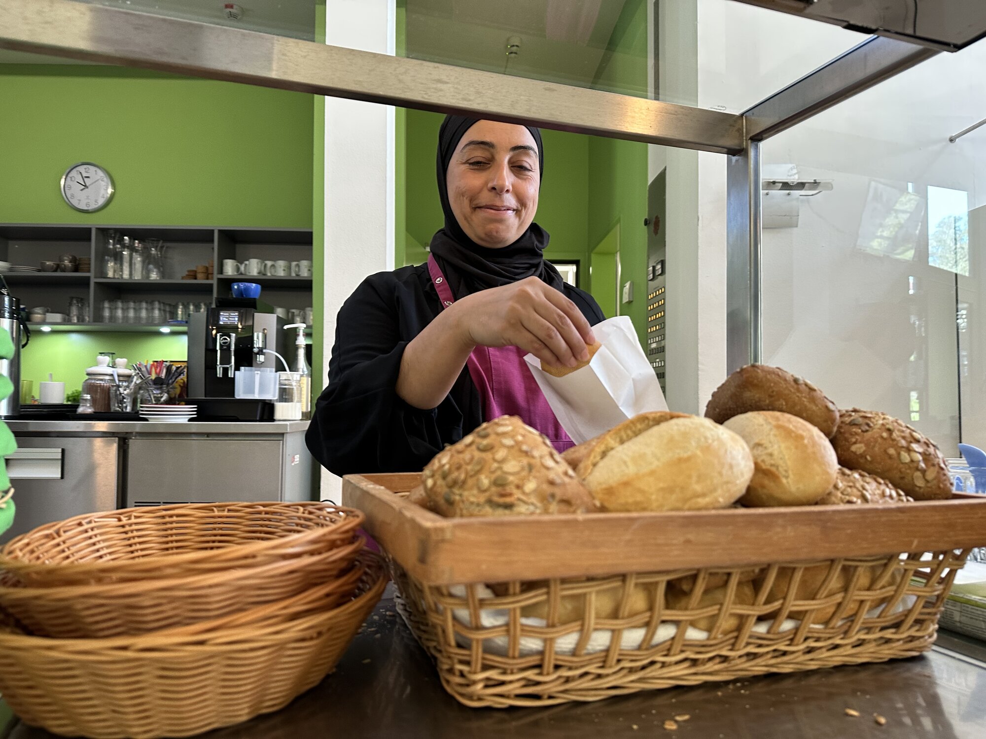 Eine Frau mit eimem Kopftuch steht hinter einer Theke und packt ein Brötchen in eine Tüte.