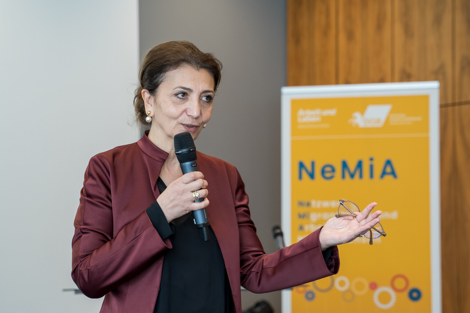 Eine Frau (Naciye Celebi-Bektas) hält ein Mikrophon in der Hand und hält eine Rede. Hinter ihr ist das Plakat des Netzwerkes NeMiA zu sehen.